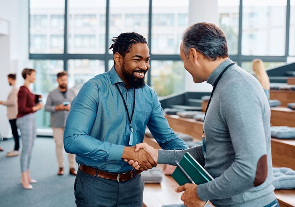 Two men are shaking hands in an casual networking environment while smiling at each other.
