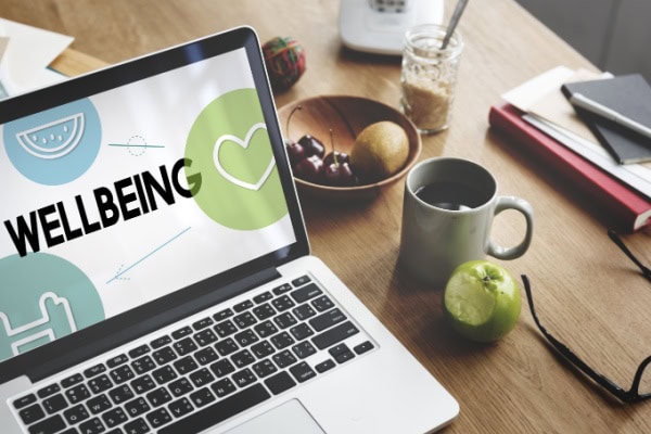 desk with laptop showing wellbeing graphic and apple coffee cup glasses and various other things on the desk