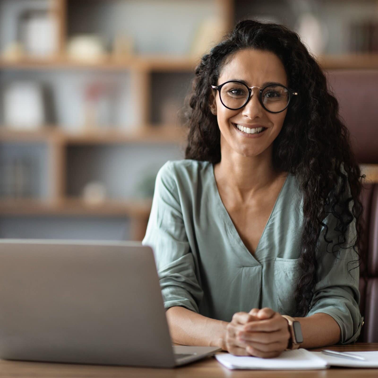 Smiling female coach educator sitting at a desk with a laptop, learning about accreditation for coach training programs.