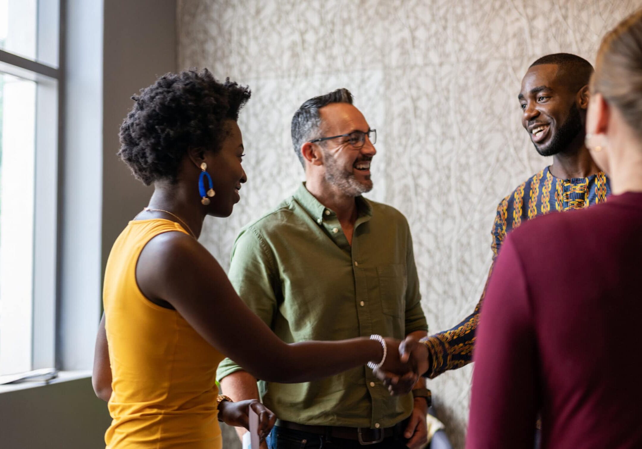 A diverse group of coaches connect warmly, shaking hands and smiling in an office setting. Two individuals greet each other in the foreground, while others stand nearby, fostering a friendly and collaborative atmosphere.