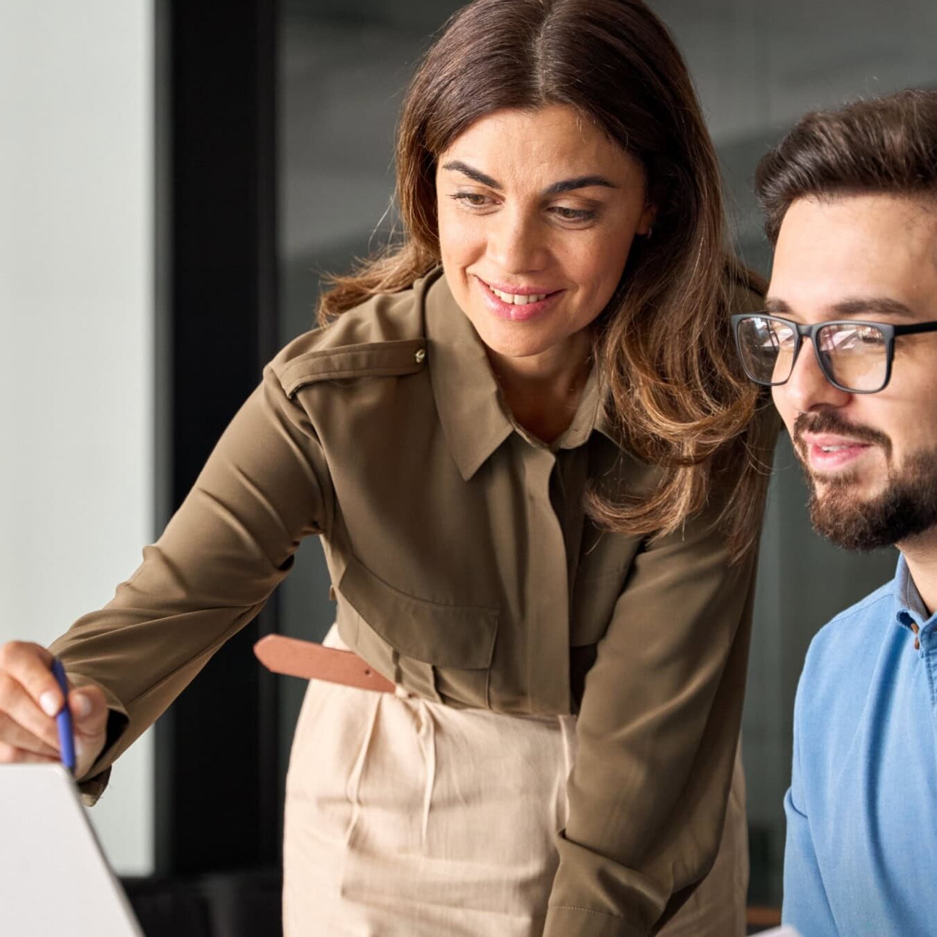 A woman helps a man on a laptop in an office setting