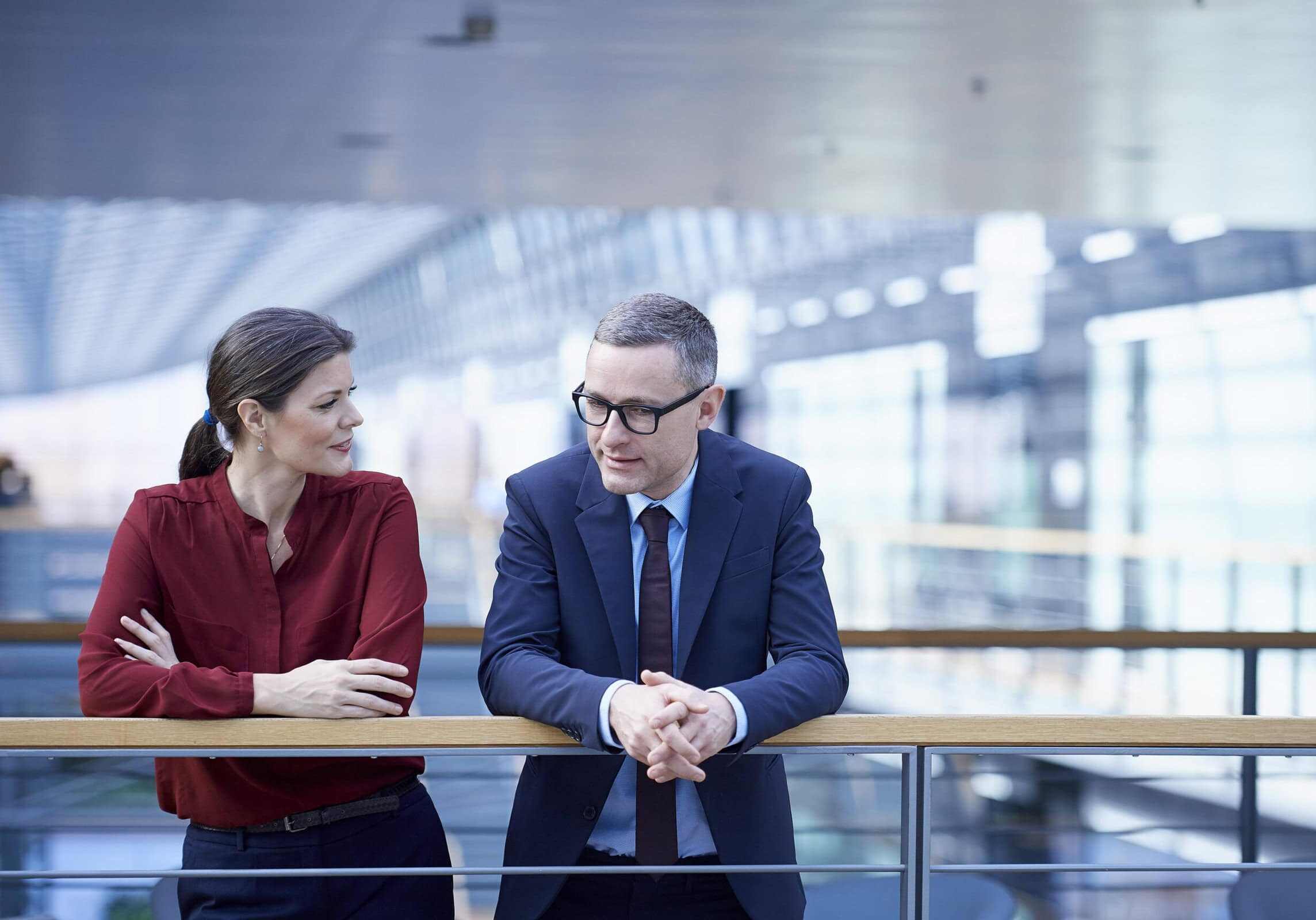 Businessman and businesswoman standing on a modern office balcony in discussion, symbolizing corporate coaching.