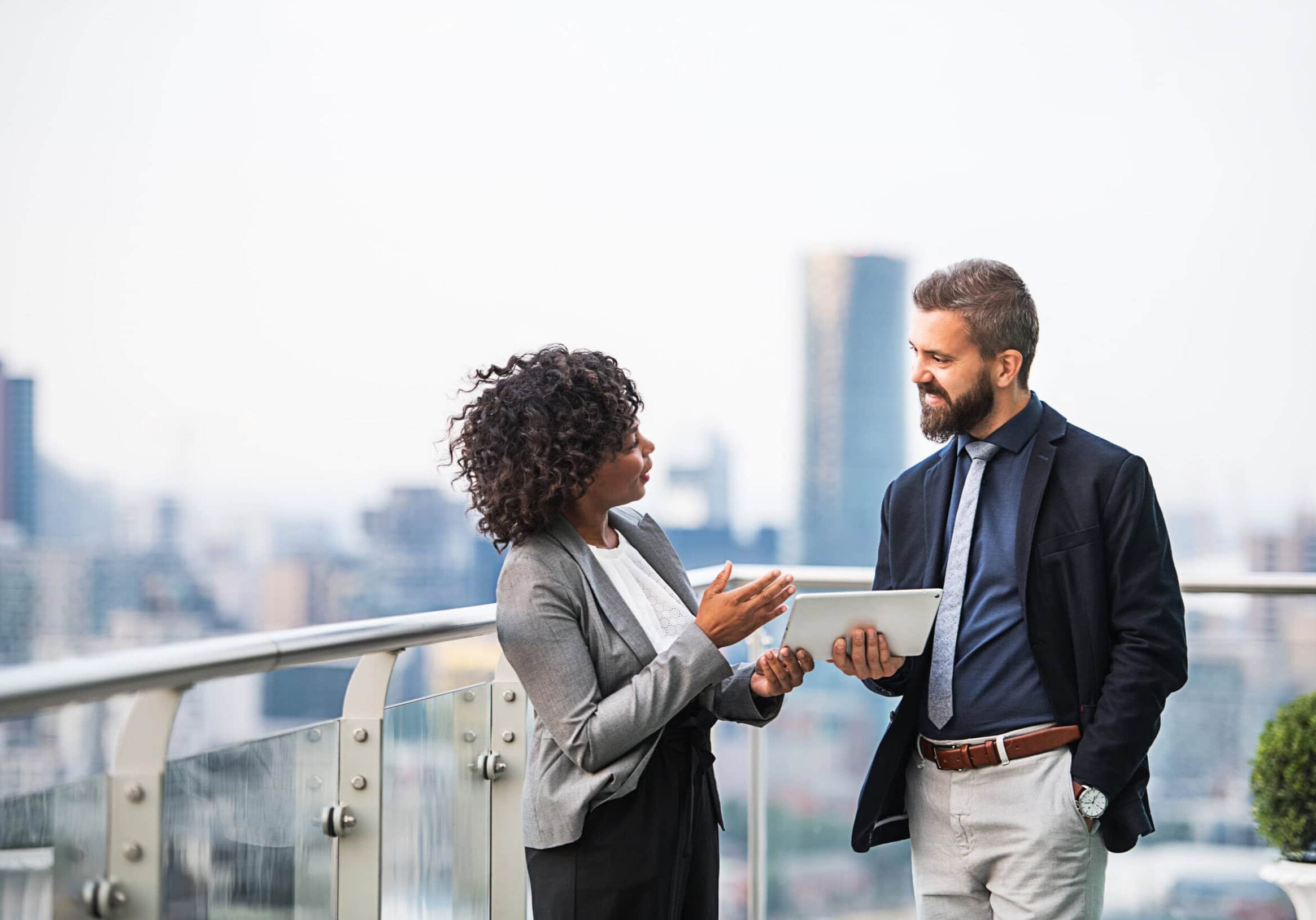 Two professional coaches having a discussion on a rooftop terrace, with a tablet in hand and a city skyline in the background.