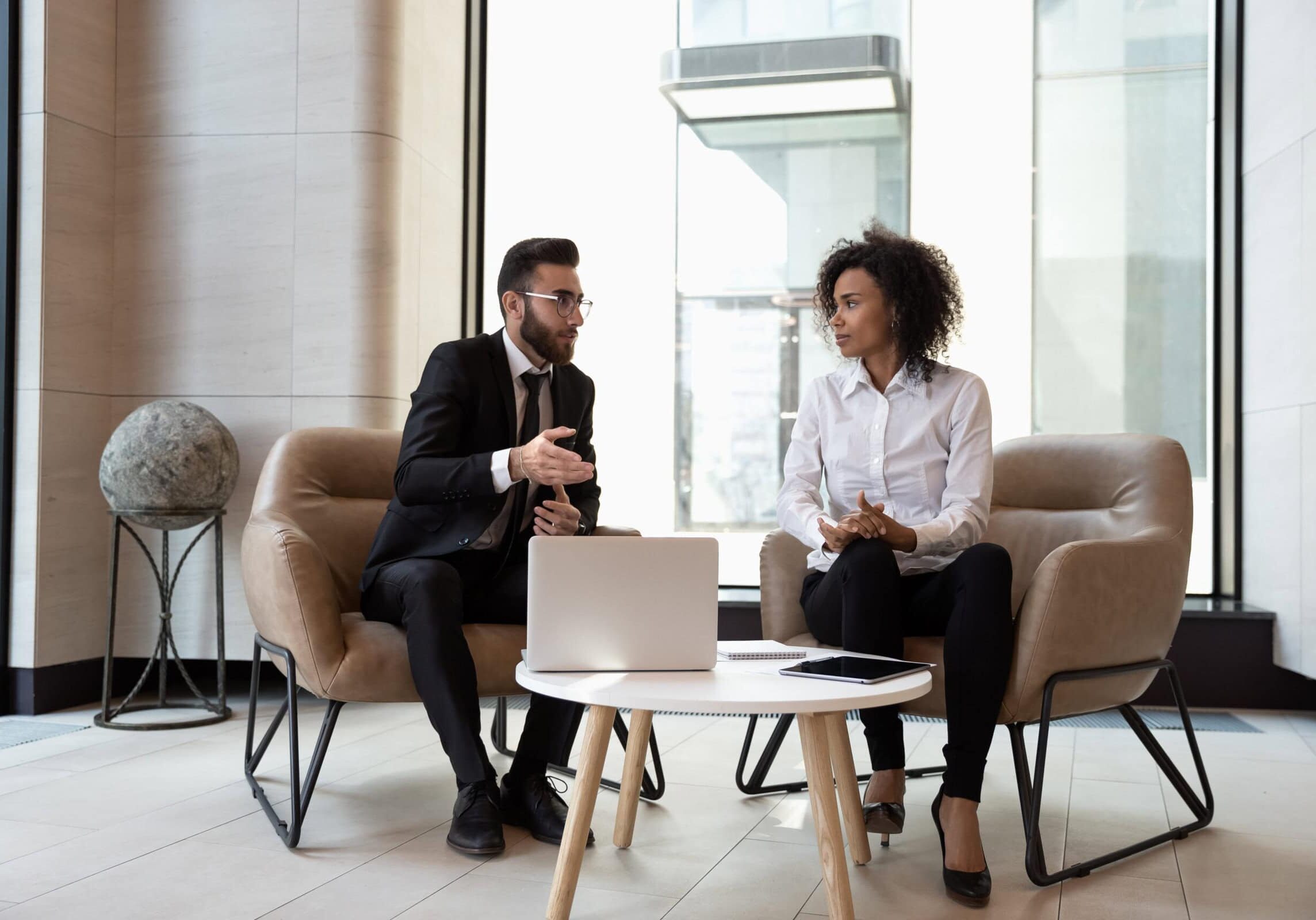 A man in a suit and a woman in professional attire seated and discussing work in a modern office setting