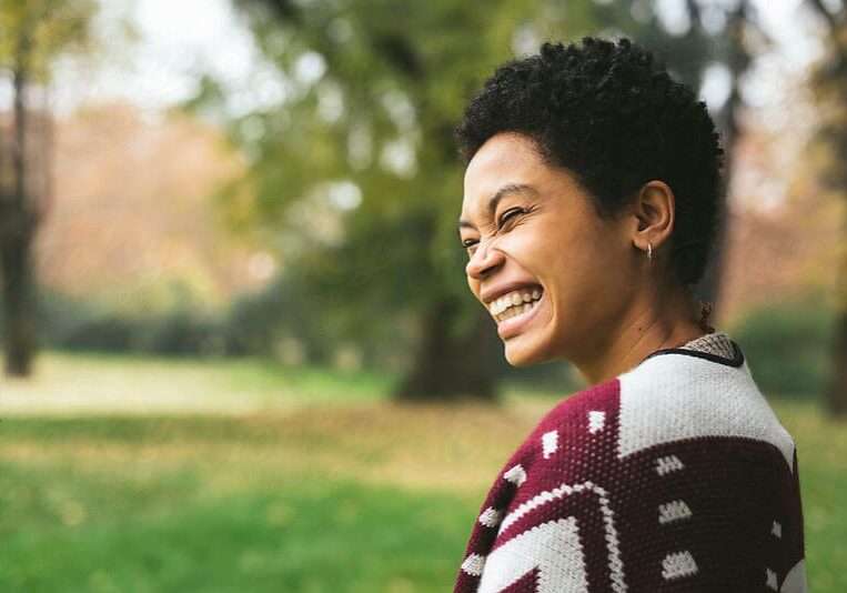 A smiling woman with short curly hair wearing a colorful patterned jacket in a natural outdoor setting, portraying positivity and joy from the coaching profession.