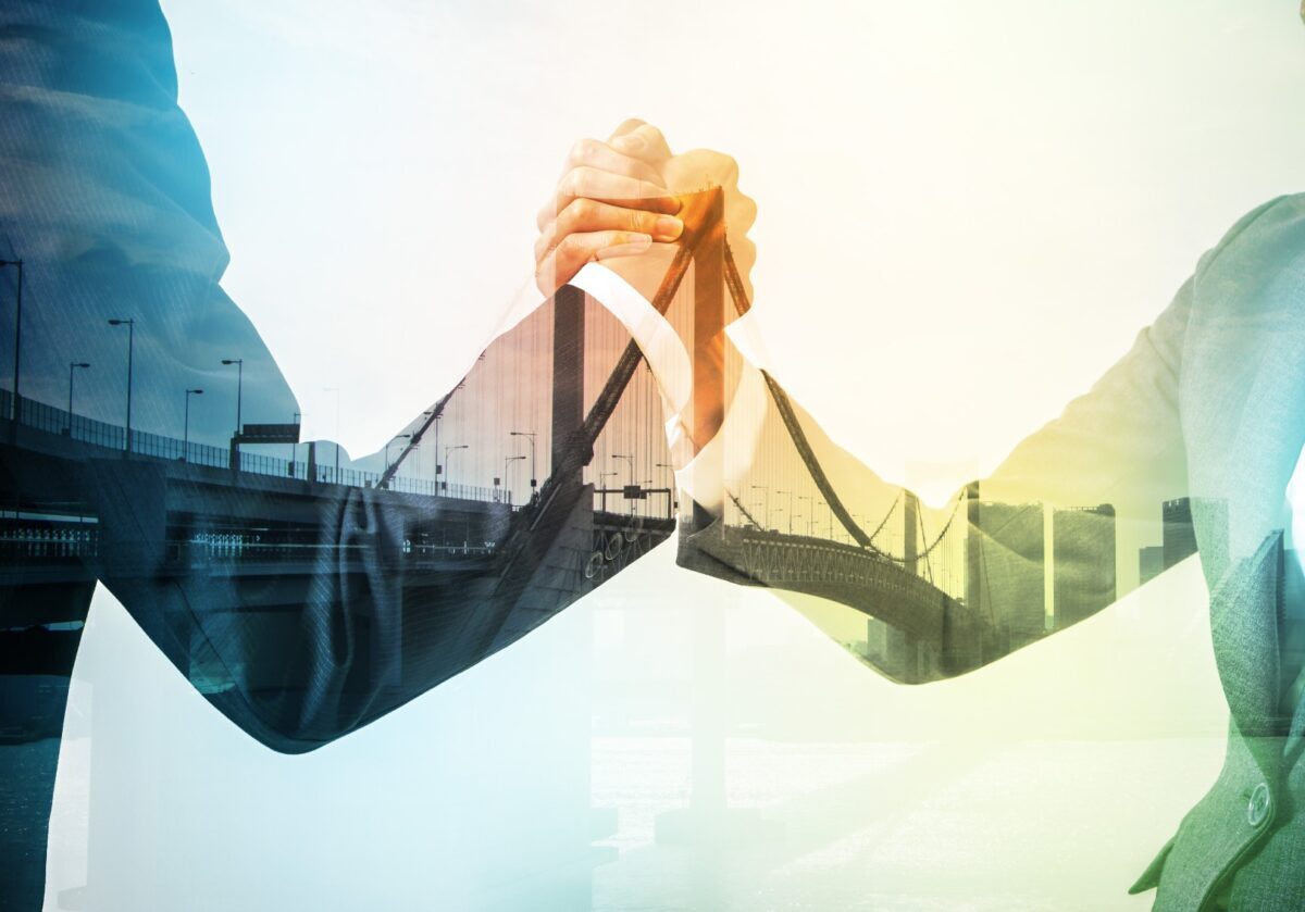 A double exposure of two business professionals clasping hands, overlaid with an image of a suspension bridge and a modern cityscape.