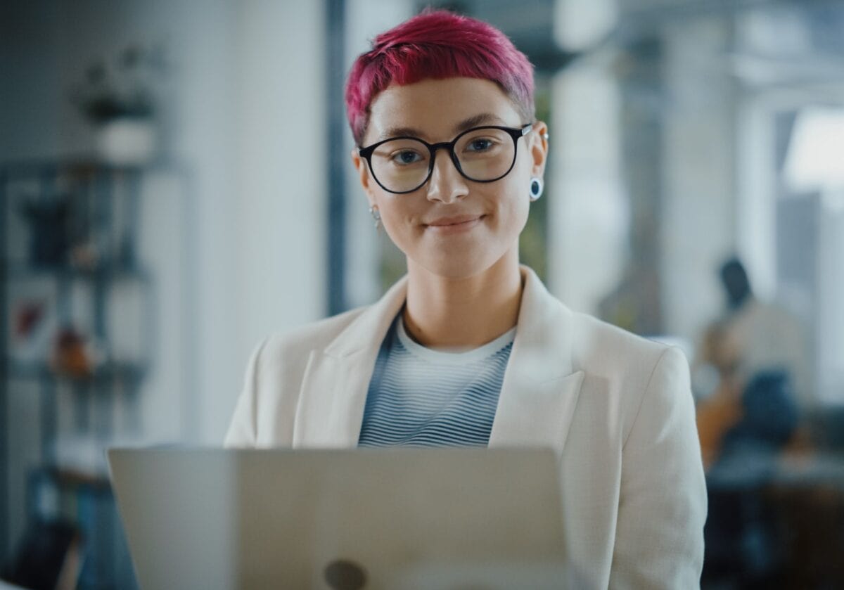   Portrait of a young, Caucasian woman with short pink hair standing, holding a laptop, looking at camera, smiling .