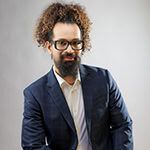 Paras Daithanka headshot photo, man with long dark hair wearing glasses and a suit smiling in front of gray background