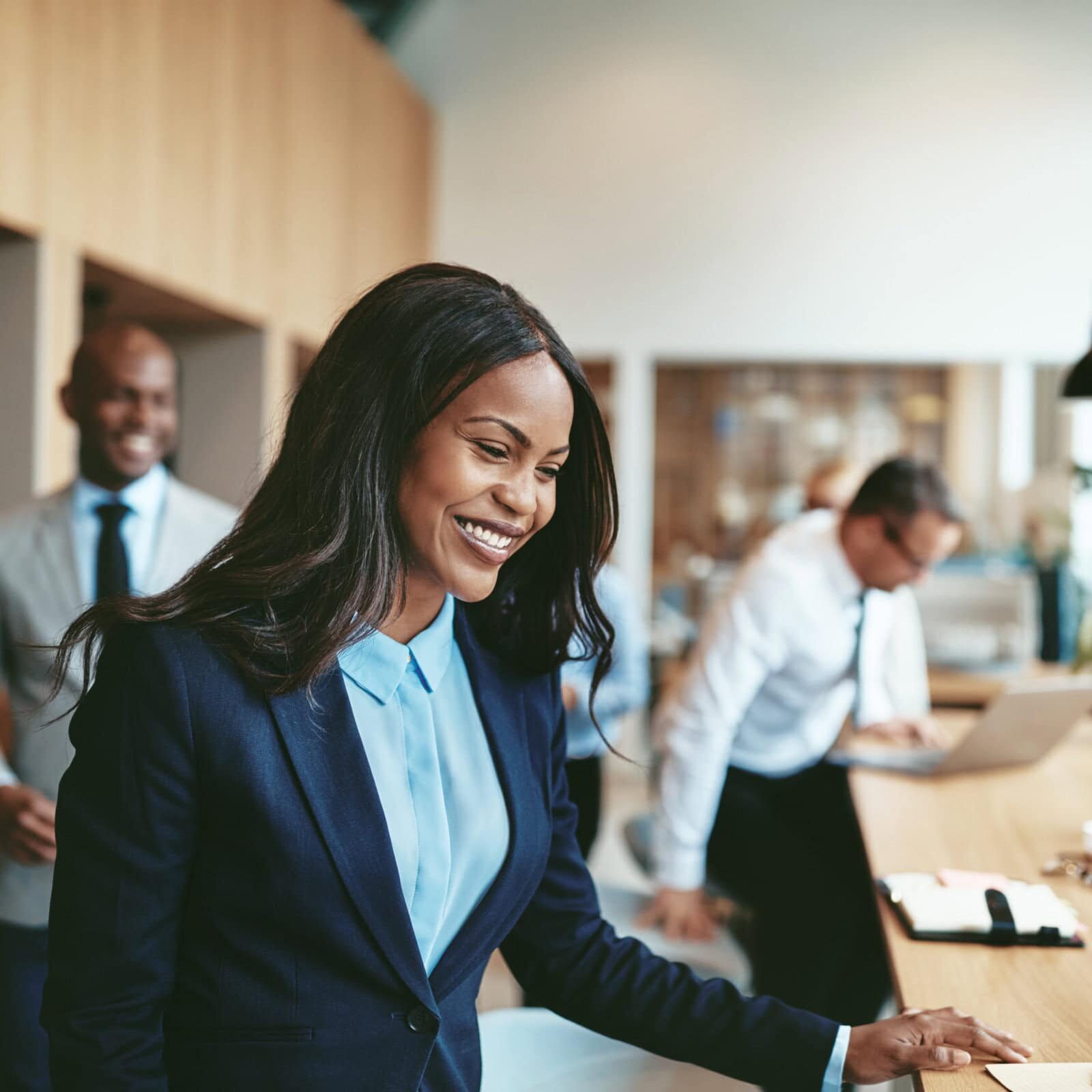 A woman is in the focal point, standing behind a desk smiling, while other co-workers are moving to sit at the same desk.