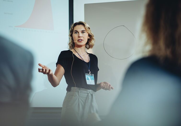 A woman is presenting in front of a group of people, with a presentation behind her.